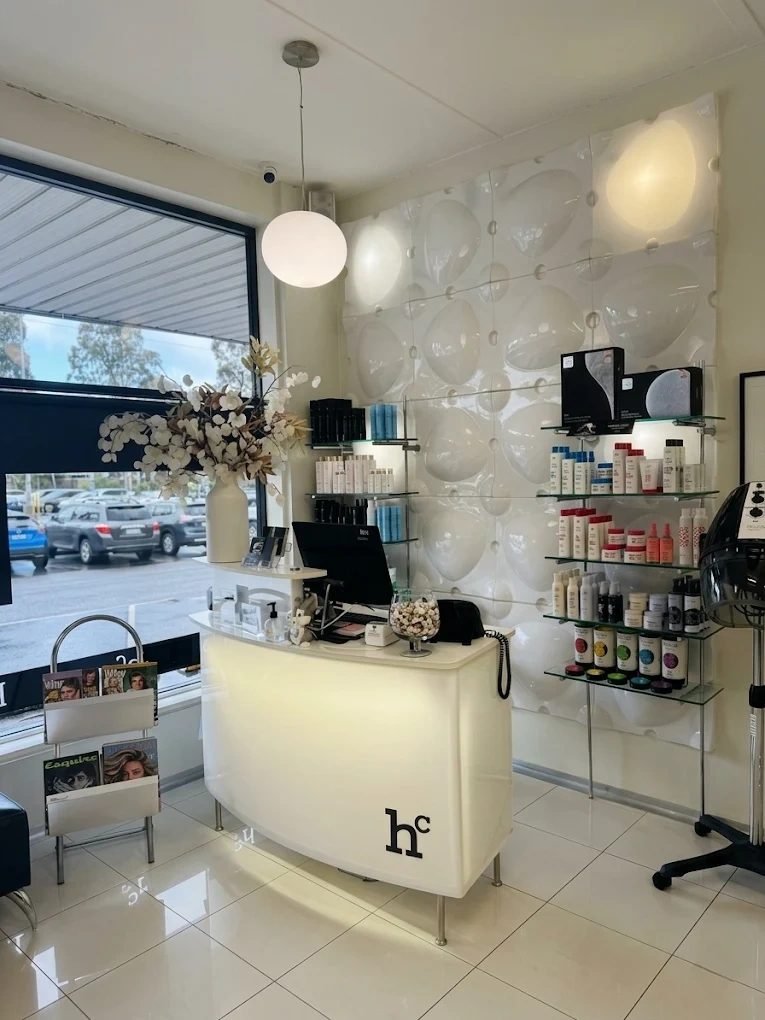 The reception area at Hallam Coiffure: a curved white desk with the hc logo, glowing white wall panels and glass shelves of hair products beside the front window