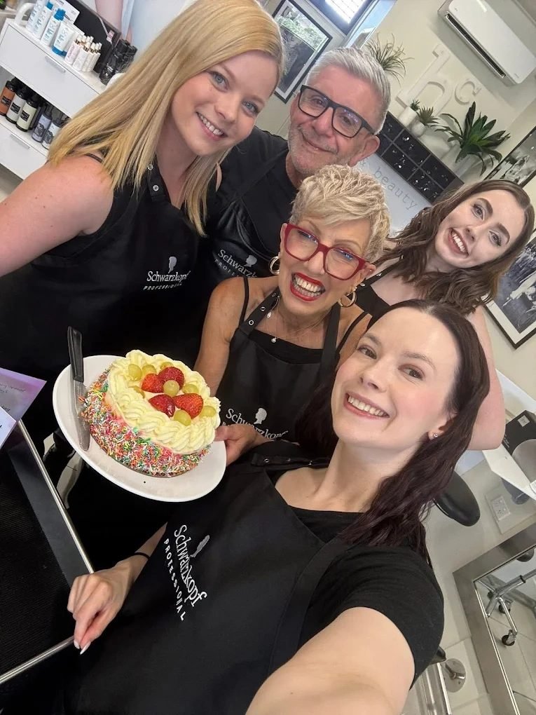 Five members of the Hallam Coiffure team in black Schwarzkopf Professional aprons, smiling around a celebration cake